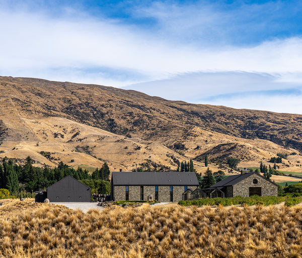 A scenic landscape photograph of a farm with stone and dark-roofed buildings nestled at the base of large dry grass-covered hills under a blue sky with light clouds.